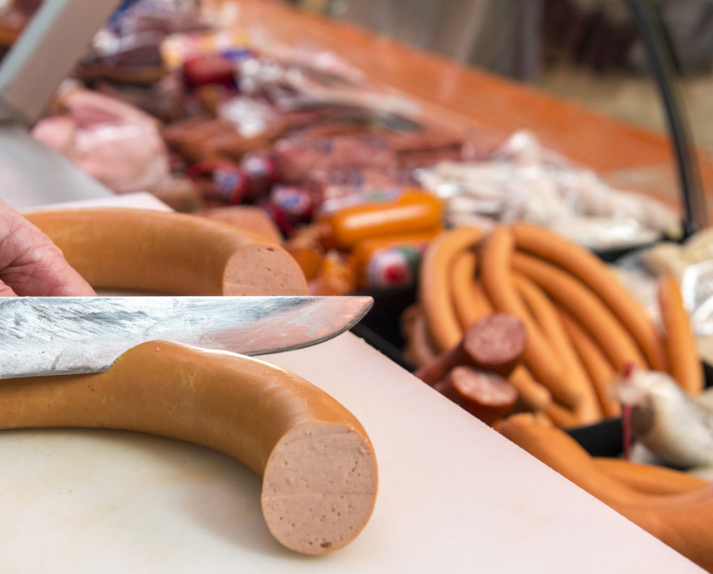 Fleischwurst wird an der Wursttheke mit Messer geschnitten. Im Hintergrund Wurstwaren